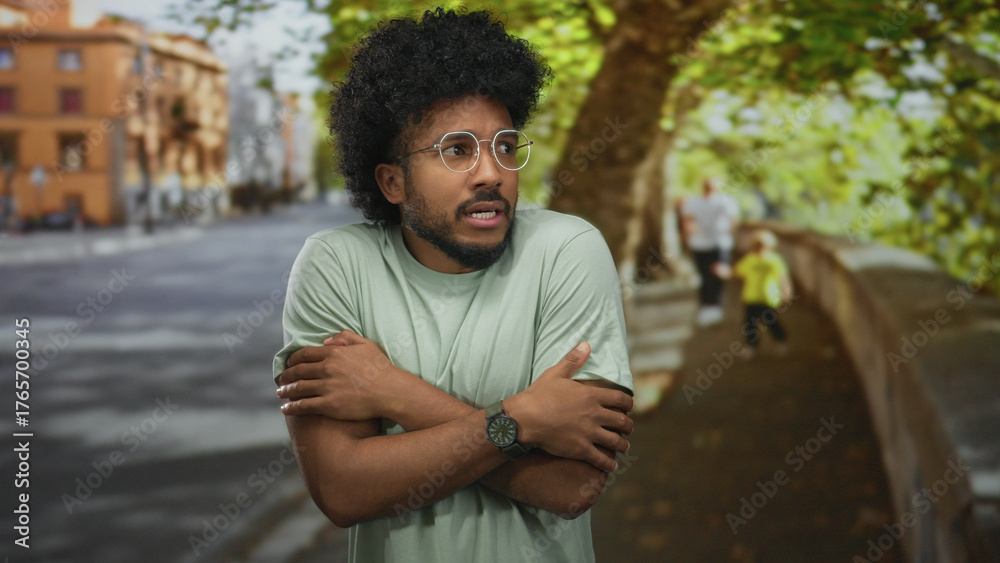 Fototapeta premium African american man with glasses nervously clasping hands on a city street with trees and buildings in the background.