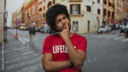 Fotografía Man in red lifeguard shirt stands thoughtfully on blurred city street with historic buildings