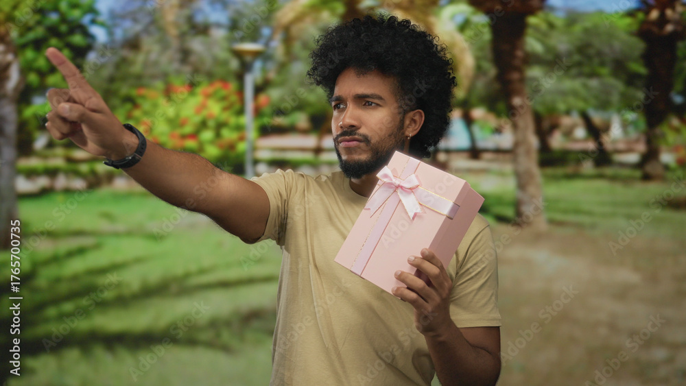 Fototapeta premium Man in park holding a pink gift box with ribbon in a green outdoor setting exhibiting a thoughtful expression highlighting nature and celebration vibes.