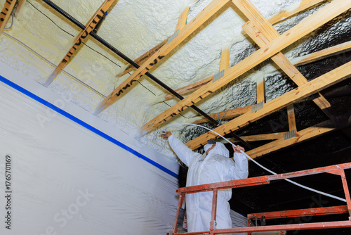 Fotografie Painter in protective gear uses spray machine to apply refractory paint insulation to foam ceiling beams of construction site