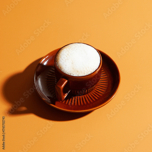 Small brown ceramic cup and saucer hold a frothy cappuccino with fine milk bubbles at the rim. Ridged saucer and strong side lighting cast a crisp lower left shadow, producing warm retro mood