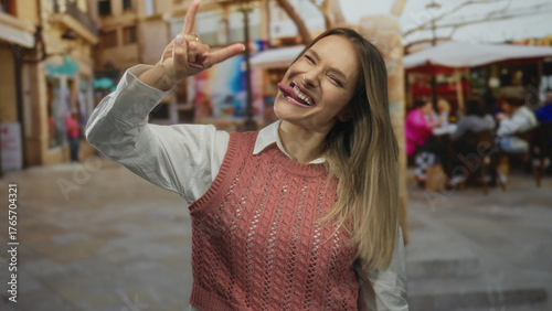 Woman smiling doing rock gesture on city terrace with people in background enjoying coffee at restaurant, wearing pink sweater, lovely day outdoors.