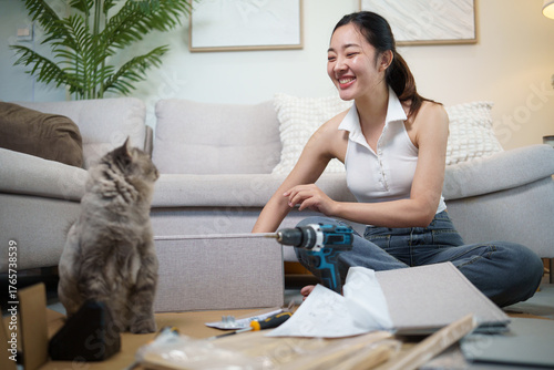 Smiling woman holding a power drill while her cat curiously watches during a DIY furniture project.
