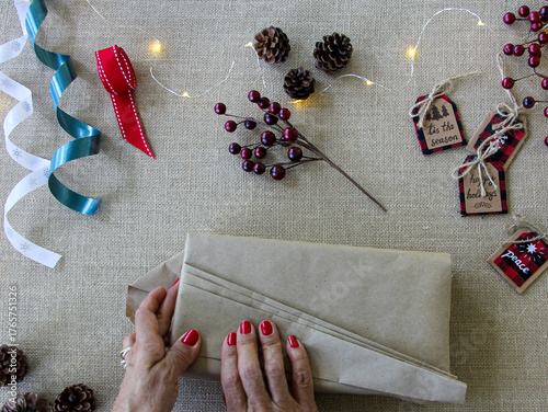 Flat lay overhead of woman wrapping Christmas gifts with Christmas supplies to pack Holiday presents on textured neutral surface using recycled kraft Paper.