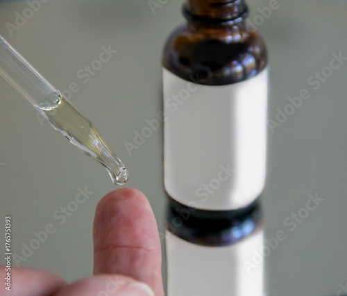 Closeup view of woman using a dropper to place a drop of serum on her fingertip and a glass serum bottle with white mockup friendly label in the background for skincare concept. 