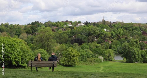 Couple sitting on a bench facing a lush valley of trees and rooftops on a bright spring day. Quiet pause, shared moment, nature and city meeting at the edge.
