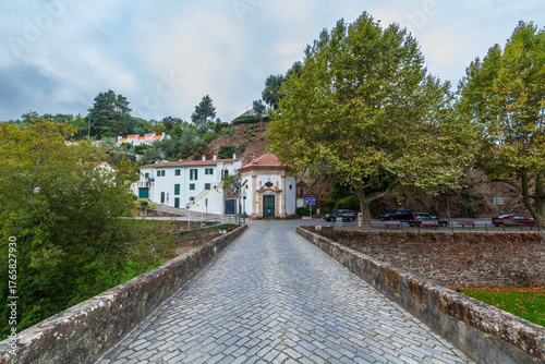 A cobblestone bridge leads toward a white chapel with a red-tiled roof