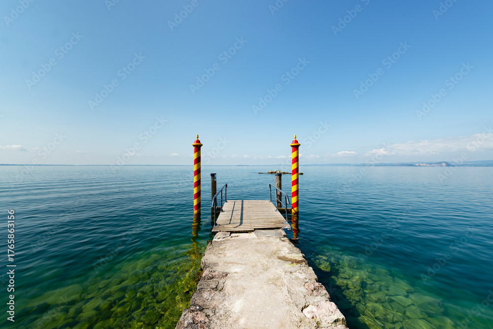 Fototapeta premium Gelb-rot gestreifte Holzstangen am Bootsanleger der Locanda San Vigilio auf einer Halbinsel im Gardasee in der Morgensonne, Venetien, Italien