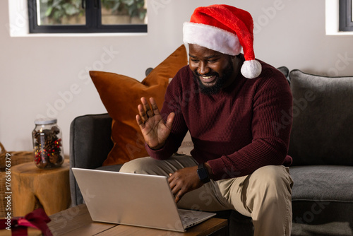 African American man waving at laptop while sitting at home near pinecone jar and Santa hat