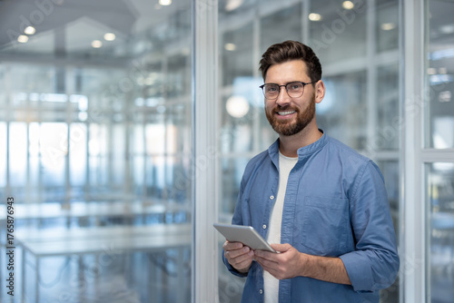Young businessman wearing glasses and a blue shirt holding a digital tablet and smiling at the camera in a modern glass office interior, symbolizing business, technology, and success
