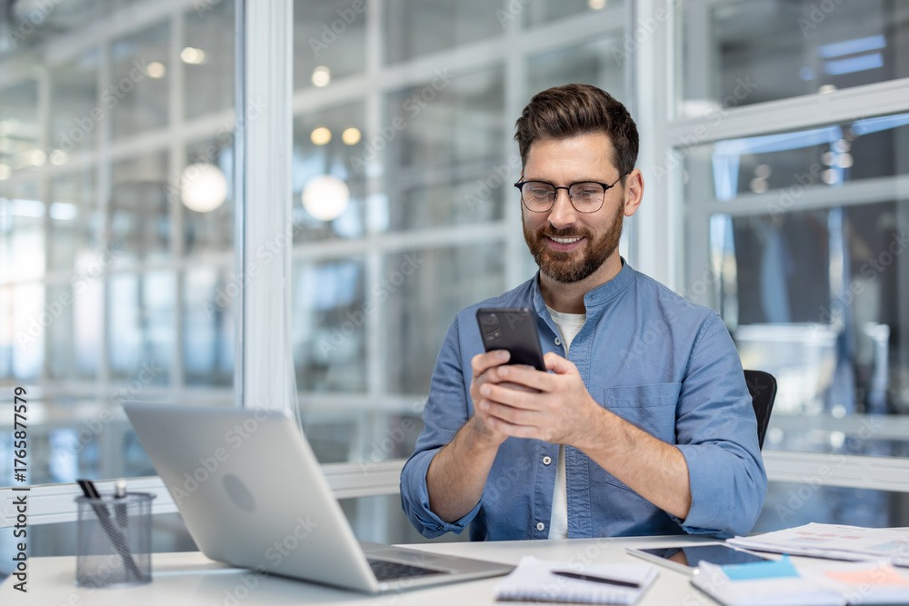 Obraz premium Smiling businessman wearing glasses and a beard sitting at a desk, looking cheerful while messaging on a smartphone, with a laptop and documents on the contemporary office table