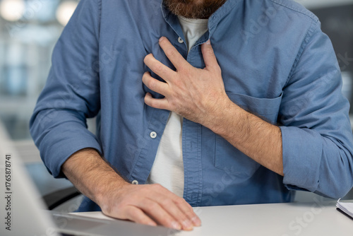 Man clutching chest at office desk, grimacing from sudden acute chest pain suggesting heart attack or severe cardiac distress during work urgent medical crisis and discomfort