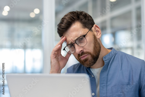 Young bearded man with glasses at a laptop, rubbing his temples and wincing from a headache while stressed, tired and overwhelmed by work and office pressure