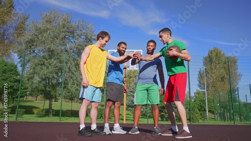 Motivated athletic diverse multiracial male basketball players with ball standing on outdoor court, making stack of hands, expressing good sportsmanship, unity and readiness for streetball game.