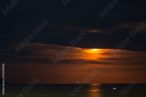 A stunning blood moon over the ocean, casting a golden-orange glow across the sky and reflecting on the sea. A small boat and distant lights add depth to the tranquil night scene.