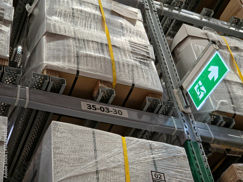 Fotografie A low-angle view of tall metal warehouse shelving stacked high with shrink-wrapped boxes, featuring a brightly lit green exit sign and rack labeling