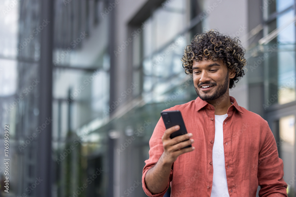 Obraz premium Close-up photo of a young Indian man standing on the street with a smile and using a mobile phone