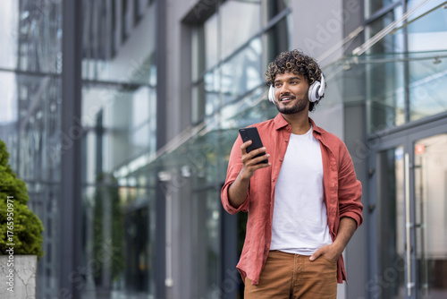 Photography Photo of a young smiling Indian man wearing headphones walking around the city,