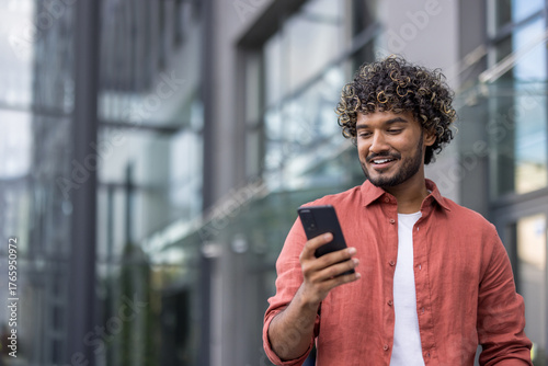 Close-up photo of a young Indian man standing on the street with a smile and using a mobile phone