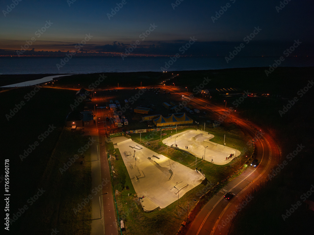 Fototapeta premium Aerial night view of skatepark brightly lit concrete ramps and bowls stand out against dark surroundings, nearby roads and vehicles, distant city lights shimmer across water under night sky.