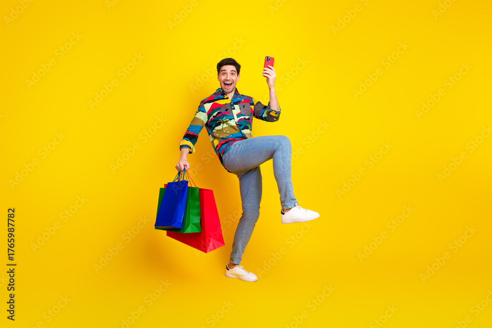Obraz premium Young man in colorful shirt jumps with shopping bags and takes a selfie against a bright yellow background expressing joy and fun shopping fashion and leisure