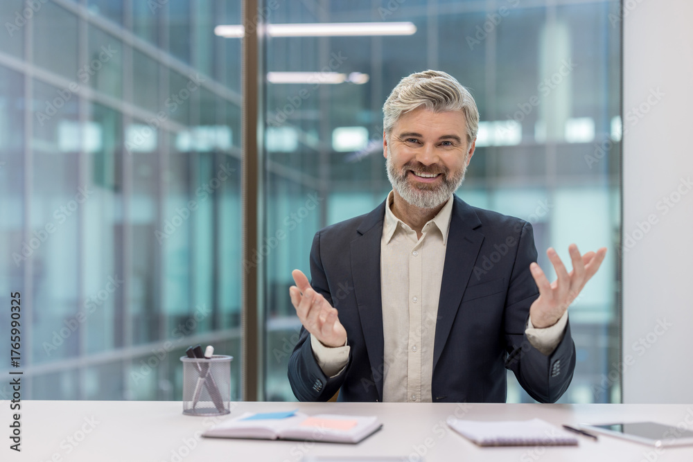 Fototapeta premium Mature executive, a confident businessman with a grey beard, smiling at the camera and gesturing with his hands during a virtual meeting or online presentation from a modern office desk