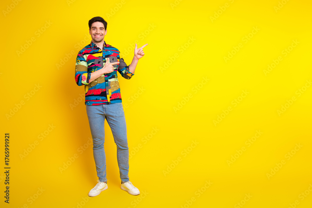 Fototapeta premium Young man in colorful shirt poses against bright yellow background smiling and pointing