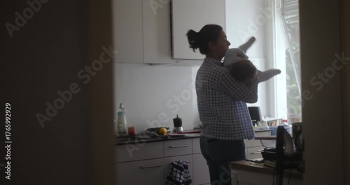 Mother holding baby in kitchen during quiet morning light, tender everyday moment showing care, warmth, and the soft rhythm of domestic life