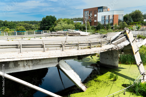 A concrete road bridge, destroyed as a result of hostilities, collapses over a ditch of stagnant green water, symbolizing the devastating impact of war