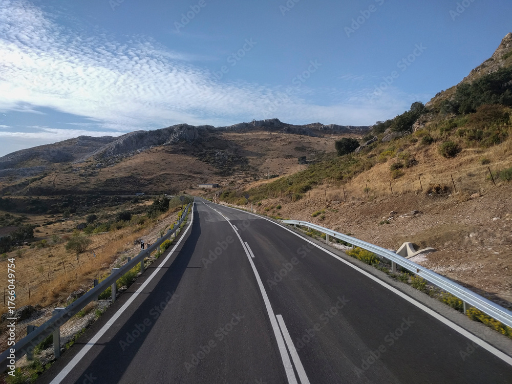 Fototapeta premium Spain - A winding asphalt road with white markings extends through a dry, mountainous landscape under a bright blue sky with scattered clouds.