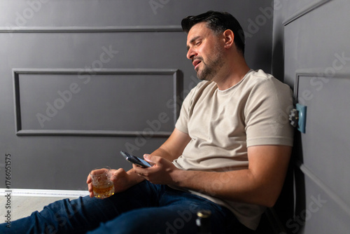 Man sitting on floor with drink, struggling to stay awake in a dimly lit room