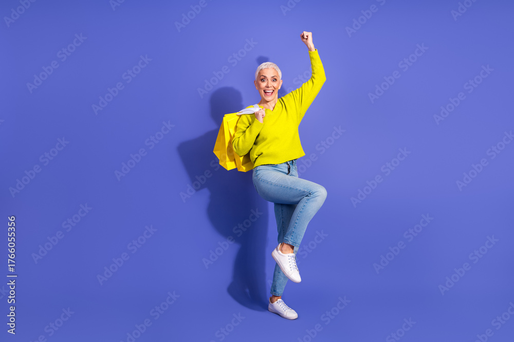 Fototapeta premium Cheerful mature woman in bright yellow sweater jumping with shopping bags against a purple backdrop ready for shopping and promotion