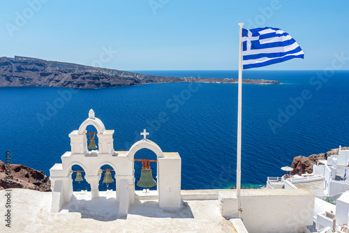 Greek orthodox church with bells and greek flag against famous white houses on Santorini island, Aegean sea, Greece.