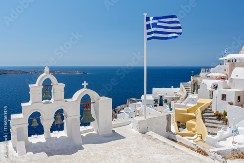 Greek orthodox church with bells and greek flag against famous white houses on Santorini island, Aegean sea, Greece.