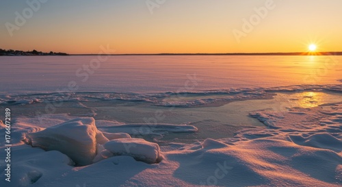 A snow-covered lake reflects the warm hues of a sunrise, breaking ice in the foreground, with distant shore