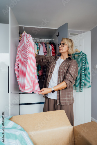Middle aged woman selecting clothes from wardrobe for donating to Charity shop. Decluttering, Sorting clothes and Cleaning Up. Reuse, second-hand concept. Conscious consumer, sustainable lifestyle.