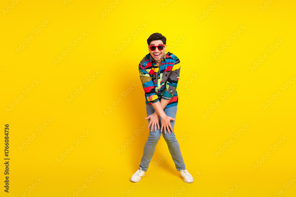 Fototapeta premium Young man in colorful patterned shirt wearing red sunglasses poses playfully against a bright yellow background for fashion lifestyle and advertising uses