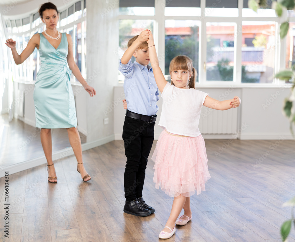 Naklejka premium Couple of positive interested preteen dancers, boy and girl preparing for performance, rehearsing ballroom dance in spacious classroom while female teacher observing from background