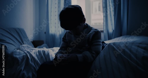 Child sitting on edge of bed in dim blue-lit room with head lowered, conveying childhood loneliness, melancholy, and silent emotional struggle