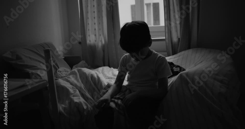 Boy sitting on bed with head lowered in quiet bedroom, appearing thoughtful and withdrawn, symbolizing sadness, loneliness, emotional struggle, and childhood solitude