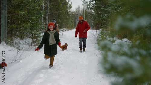 Wide shot of active young Caucasian parents and children in warm winter coats running and playing in snow, while walking outdoors in forest during vacation, small pet dog jumping around