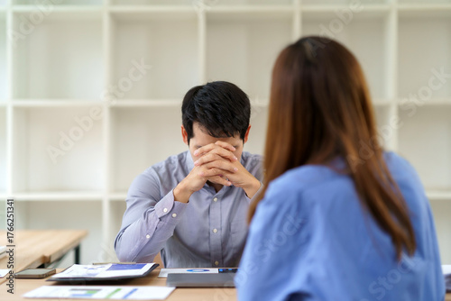 Stressed man experiencing symptoms of burnout, sitting at a desk with his hands clasped, facing a colleague in an office meeting