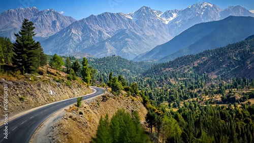 Winding road along a mountain valley with forests and snow-capped peaks on a sunny day
