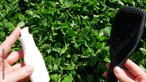 Female Hands Holding Hair Product and Comb on Organic Green Grass Background, Copy Space