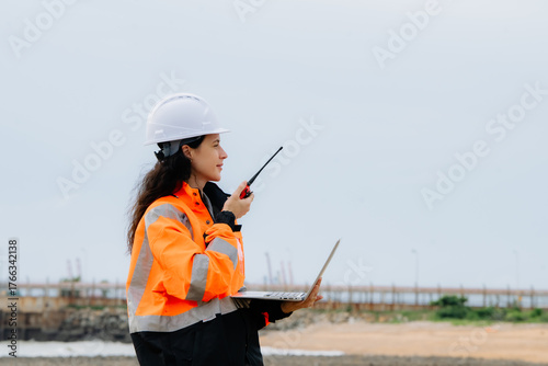 Slika na platnu A woman wearing an orange jacket and a white helmet is holding a walkie talkie