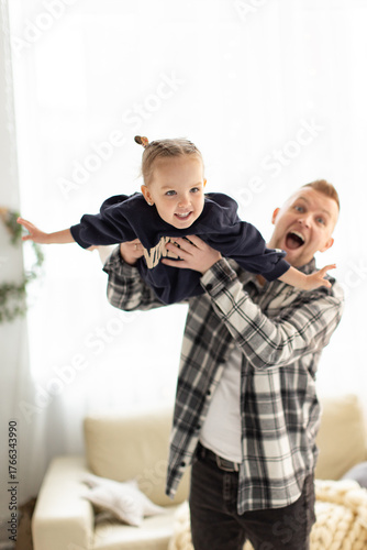 A happy father holds his young daughter up in the air, both smiling and enjoying a playful moment indoors.