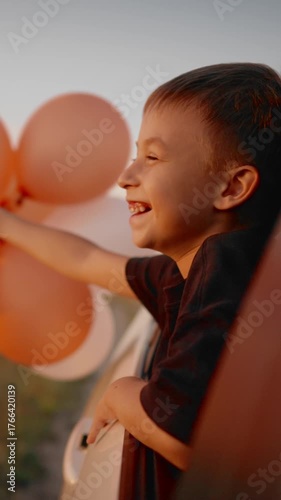Vertical portrait of cheerful boy enjoying summer wind from car window, balloons for birthday party in hands swaying by air stream. Carefree childhood and freedom, happy smiling child feeling joy