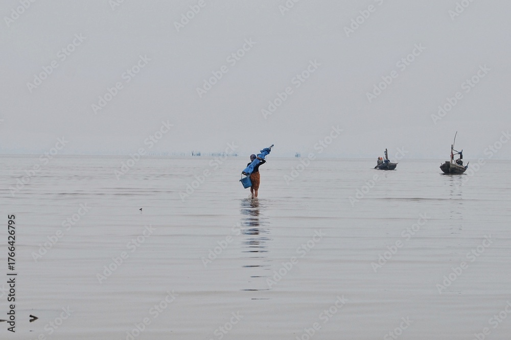 Naklejka premium Fisherman Wading in Shallow Sea with Small Boats in Misty Horizon Under Gray Sky
