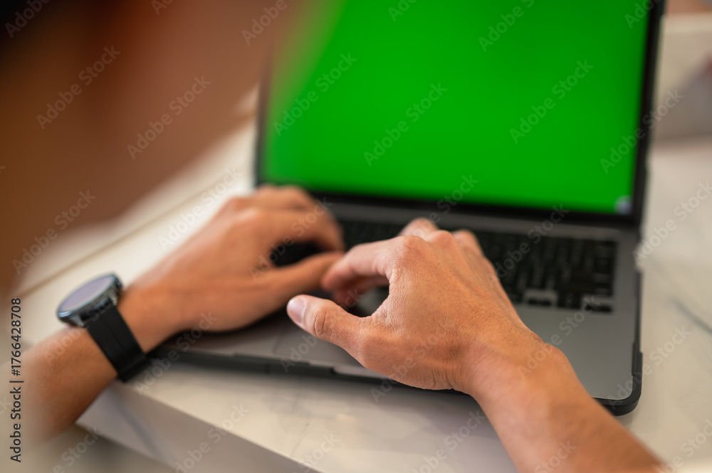 Fototapeta premium Close-up of an Asian man’s hands typing on a laptop with a green screen at a café table, showing modern workspace setup and technology focus in a professional environment