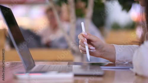 Woman is writing on a laptop with a white pen. She is sitting at a table. There are other people in the background